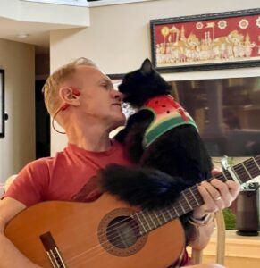 A black cat sitting on Karl's guitar as he plays the guitar.