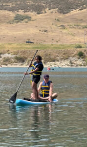 Miti standing on a paddleboard paddling while Karl sits on the paddle board.