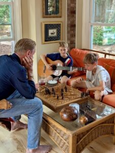 Karl playing chess against a young boy while another young boy strums on the guitar.