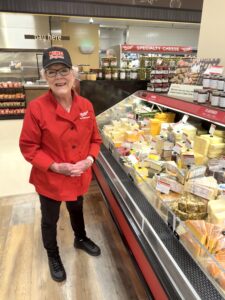 A lady wearing a red shirt and a black hat at the cheese counter of a grocery store in Colorado.