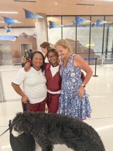 Three people posing together for a photo at the airport as a big black dog sniffs the suitcase.