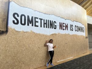 Mitia standing in front of a sign that says "Something new is coming." 