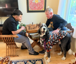 Two gentlemen playing chess with Plato the big dog nearby.