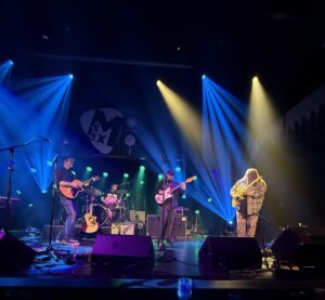 Three men on stage playing guitars and one man playing the drums.