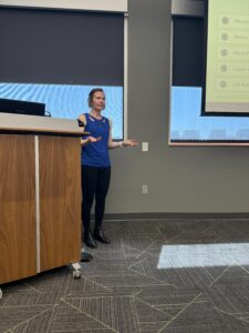 A woman in a blue shirt and black pants speaking to a group of people.